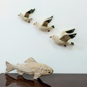 May include: Three white felt seagulls with black and green wings are flying in formation on a white wall. A wooden fish sculpture is on a dark brown surface below the seagulls.