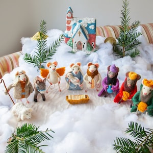Könnte beinhalten: Eine skurrile Weihnachtskrippe mit Filz-Mausfiguren. Die Szene umfasst eine Kirche, ein Schaf, einen Esel, einen Engel und Figuren, die die Weihnachtsgeschichte darstellen. Die Figuren sind auf einer schneebedeckten Landschaft mit Tannengrün-Akzenten angeordnet.