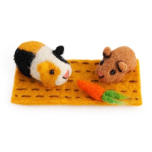 Puede incluir: Una escena de fieltro con dos cobayas y una zanahoria. Una cobaya es tricolor con pelaje negro, blanco y naranja. La otra es marrón. Están sobre una estera rectangular amarilla, con una zanahoria naranja con hojas verdes.