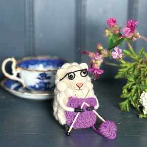 May include: A white felt sheep wearing glasses and knitting a purple scarf with two knitting needles.