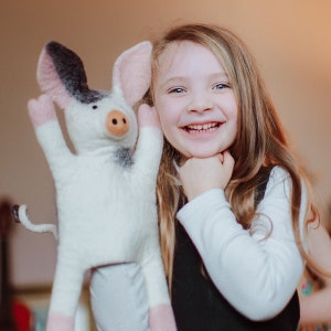 May include: A young girl with long blonde hair smiles while holding a white and black felt pig puppet with pink ears and hooves.