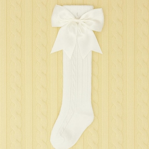 May include: White knee-high socks with a large satin bow at the top. The socks have a cable knit pattern and are displayed against a light yellow background with a textured design. A classic accessory for children.