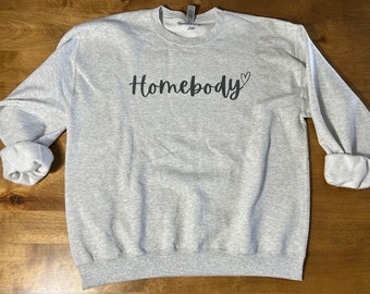Puede incluir: Una sudadera gris con el texto "Homebody" en letras negras y un pequeño símbolo de corazón.