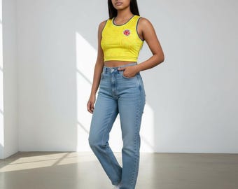 May include: A woman wearing a yellow crop top with a blue trim and a red emblem, paired with light blue jeans and white sneakers. The top has a subtle pattern. The outfit is casual and modern.