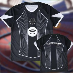 Puede incluir: Camiseta de fútbol en blanco y negro con cuello en V y detalles blancos en las mangas. La camiseta presenta un logotipo de Spotify y un escudo del equipo en la parte delantera. La parte trasera de la camiseta tiene el texto "NAME HERE".
