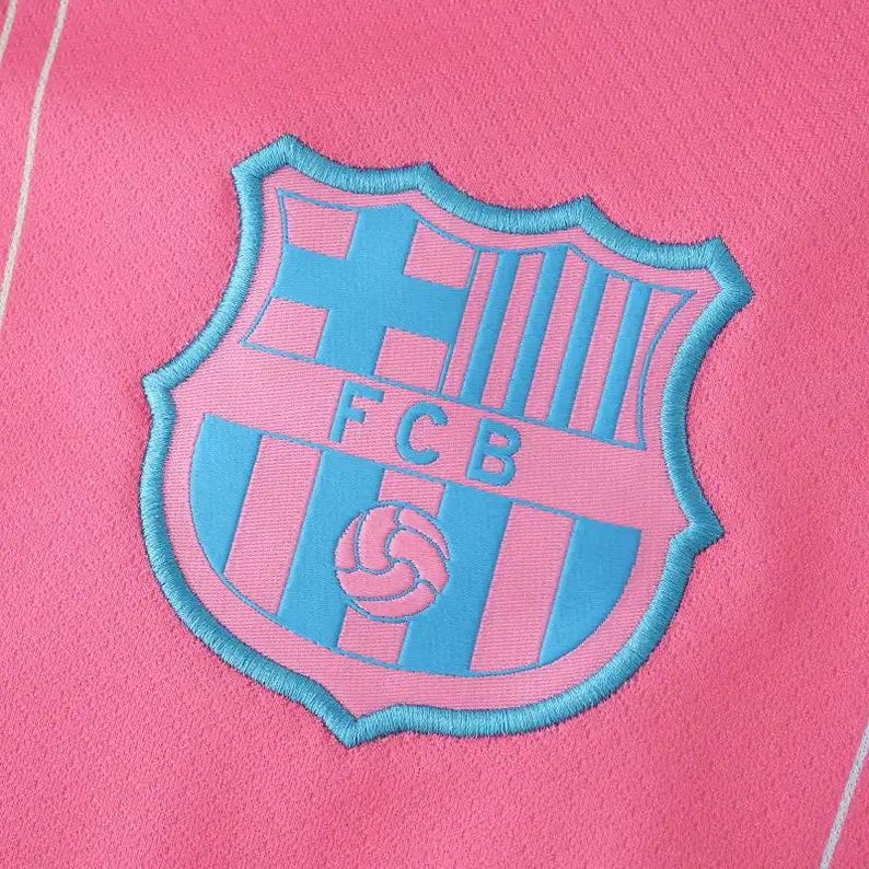 K&ouml;nnte beinhalten: Nahaufnahme eines pinkfarbenen Stoffes mit einem hellblauen Emblem. Das Emblem zeigt ein Schilddesign mit den Buchstaben "FCB" und einem Fu&szlig;ball. Das Schild ist hellblau umrandet.