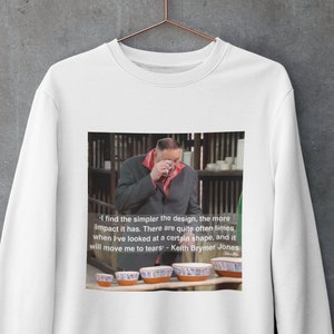 Puede incluir: Sudadera blanca con una imagen impresa de un hombre llorando, con la cita: "Cuanto más sencillo es el diseño, más impacto tiene...me hará llorar." Debajo hay varios cuencos de cerámica.