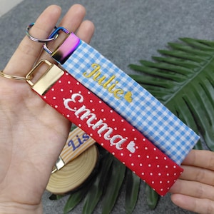 May include: Two fabric keychains with metal hardware. One is blue and white gingham with the embroidered name Julie in gold. The other is red with white polka dots and the embroidered name Emma in white. Both feature a small heart icon.
