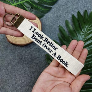 May include: A white canvas keychain with a black embroidered saying: "I Look Better Bent Over A Book".
