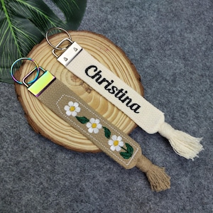 Op de afbeelding: Twee sleutelhangers met een kwast. De ene sleutelhanger is beige met de naam Christina geborduurd in zwart. De andere sleutelhanger is beige met een wit madeliefjespatroon.