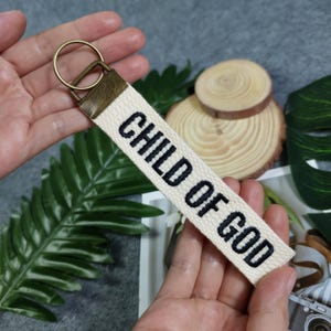Może przedstawiać: Biały brelok z płótna z haftowanym czarnym napisem "CHILD OF GOD" i złotym metalowym kółkiem na klucze.