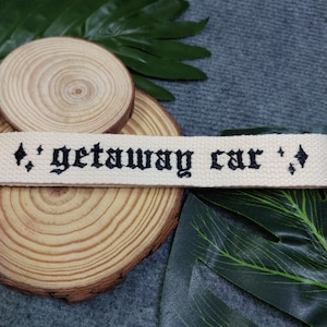 May include: A white cotton webbing keychain with black embroidered text that reads "getaway car" with diamond symbols on either side.