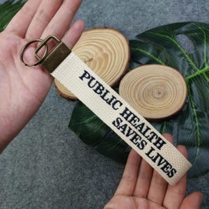 Puede incluir: Un llavero de lona blanco con un anillo de llave de latón y el texto "Public Health Saves Lives" bordado en negro.