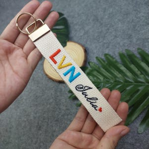 May include: A white canvas keychain with a gold key ring. The keychain has the letters "LVN" embroidered in rainbow colors and the name "Julia" embroidered in black below.
