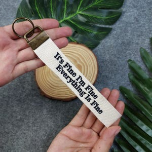 May include: White canvas keychain with a black embroidered message that reads "It's Fine I'm Fine Everything Is Fine".