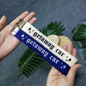 May include: Two fabric keychains with the text "getaway car" embroidered in black. One keychain is white with silver glitter and the other is blue with silver glitter.