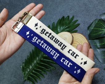 May include: Two fabric keychains with the text "getaway car" embroidered in black. One keychain is white with silver glitter and the other is blue with silver glitter.
