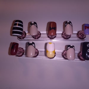 May include: A set of ten artificial nails in various designs. The nails feature patterns like black and white stripes, bows, and geometric shapes. Colors include black, white, beige, brown, yellow, and purple. The nails are displayed on a clear stand.
