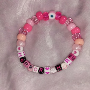 Op de afbeelding: Een ronde kralenarmband met een mix van roze, perzikkleurige en doorzichtige kralen, inclusief een centrale kraal met een boze oog-motief. De armband bevat witte letterkralen die de tekst HORROR WHORE vormen, afgewisseld met roze hartjes en smileys.