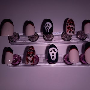 May include: A set of ten artificial nails with a Halloween theme. The nails feature a variety of designs, including a black and white ghost face, blood splatter, and a black tip with a red accent. The nails are displayed on a clear stand.