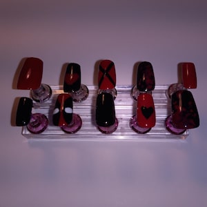 May include: A set of ten artificial nails in various designs. The nails are displayed on a clear plastic stand with purple accents. Designs include solid red, black with a red heart, and black with a red X.