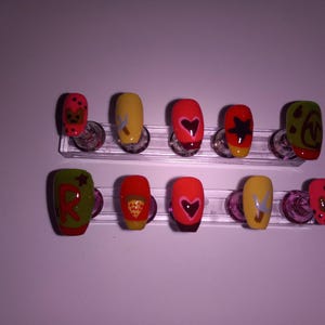 Puede incluir: Un conjunto de diez uñas artificiales, cada una con un diseño único. Las uñas presentan varios colores, incluyendo rojo, amarillo y verde, y están adornadas con corazones, estrellas y otros patrones. Las uñas se exhiben en soportes de plástico transparente.