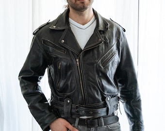 May include: Black leather biker jacket featuring multiple zip pockets, a belted waist, and white stitching. The jacket is paired with a white V-neck top and grey jeans. A classic, rugged design.