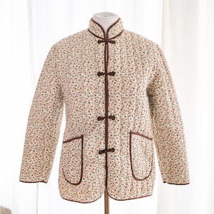 Puede incluir: Una chaqueta acolchada blanca con estampado floral y cierres de botones marrones. La chaqueta tiene un cuello mandarín y dos bolsillos.