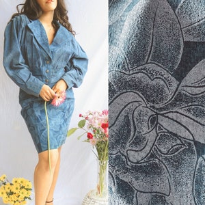 May include: A blue denim dress with a floral pattern. The dress has a collared neckline, long sleeves, and a fitted silhouette. The model is wearing the dress with a pair of brown heels.