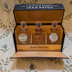 Pode incluir: Um conjunto de perfumes vintage Jean Patou numa caixa preta e bege. A caixa está aberta, revelando três frascos de perfume de vidro com tampas de cristal. Os frascos estão rotulados como "Lasso" e "Amour Amour". A caixa diz "COFFRET 3 BOULES" e "JEAN PATOU".