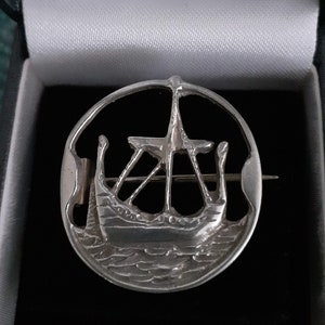 May include: A silver brooch in the shape of a Viking ship with a detailed design. The ship is sailing on a wavy sea.