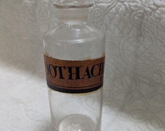 May include: Clear glass apothecary bottle with a rectangular stopper. The bottle has a brown label with the word "Toothache" printed on it. The bottle is cylindrical with a rounded base.