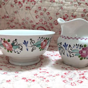 Può includere: Un set di ciotola e lattiera in ceramica bianca. La ciotola è rotonda con una base a piedistallo. Entrambi i pezzi sono decorati con motivi floreali rosa, blu e verdi e volute nere. La lattiera ha un manico e un beccuccio.