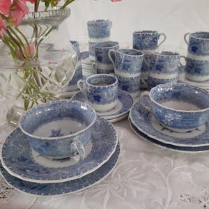 Op de afbeelding: Een verzameling blauw-witte theekopjes, schotels en mokken met bloemmotieven. De set bevat verschillende maten kopjes en schotels, gestapeld en gerangschikt op een wit kanten tafelkleed. Een heldere glazen vaas met roze bloemen staat op de achtergrond.