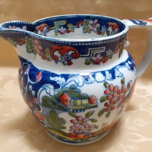 Puede incluir: Una jarra de cerámica blanca con un patrón floral azul y blanco. La jarra tiene un asa y un pico. La jarra está decorada con un patrón floral en tonos azules, rojos y verdes.