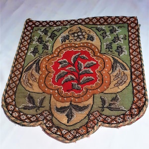 May include: A vintage, hand-stitched textile with a floral design. The central motif is a red flower with dark leaves, surrounded by a tan and green background. The border features a geometric pattern. The piece is likely a decorative item or a small bag.