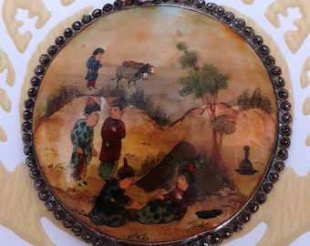 May include: A round, silver-toned pendant with a painted scene of people and animals. The scene is set in a desert landscape with a small donkey, a tree, and several figures in traditional clothing. The pendant is decorated with a beaded border.