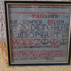 以下が含まれることがあります： 黒いフレームと金の縁取りのある額装されたサンプラー。サンプラーはリネン製で、クロスステッチのアルファベット、数字、シンボルが施されています。下部のテキストには「Margaret Hope 1819」とあります。