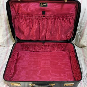 May include: An open black suitcase with a vibrant pink satin interior lining. The interior features gathered fabric pockets along the sides and a black label with gold text reading Harrods London centred at the top. It has gold-tone metal latches.