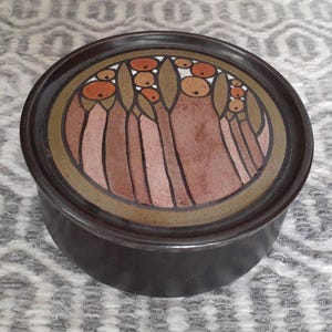 Peut inclure: Un récipient rond en céramique brun foncé avec un couvercle décoratif. Le couvercle présente un motif de formes stylisées marron et orange sur un fond brun clair, encadré d'une bordure dorée et marron.