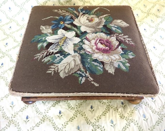 May include: A square footstool with a dark brown fabric top featuring a floral needlepoint design in white, pink, blue, and green. The edges are trimmed with a light brown rope, and it rests on four wooden feet.
