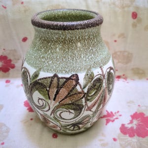 May include: A small ceramic vase with a speckled green and brown exterior. The vase has a brown rim and a white band with a floral design in brown and green. The vase is rounded in shape.