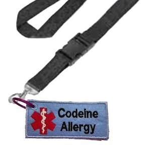 Puede incluir: Un cordón negro con un clip de plástico y una etiqueta de tela azul con un símbolo médico rojo y el texto "Codeine Allergy".