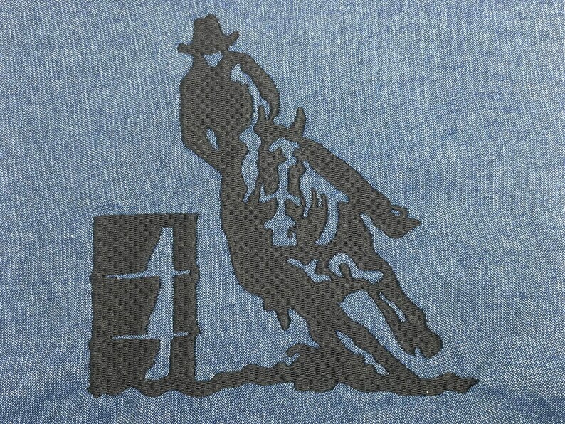 Op de afbeelding: Zwarte silhouet van een persoon die een paard over een vat rijdt, geborduurd op een blauwe denim stof.