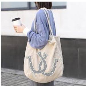 Op de afbeelding: Een grote beige tote bag met twee grijze hoefijzerontwerpen. De tas heeft een lange band en is gemaakt van een zachte, gestructureerde stof.