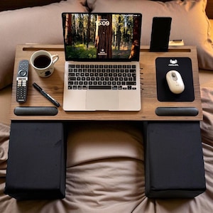Puede incluir: Un escritorio de madera para portátil con alfombrilla de ratón, soporte para teléfono y reposamuñecas integrados. Cuenta con un portátil, una taza de café, un mando a distancia y un bolígrafo. El escritorio tiene cojines negros para soporte de piernas.