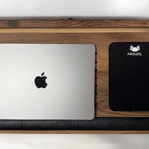 Może przedstawiać: Drewniany stojak na laptopa z czarnym sk&oacute;rzanym podparciem nadgarstka, srebrny laptop z logo Apple i czarny notes z napisem "AXOLOTL" nadrukowanym na biało.