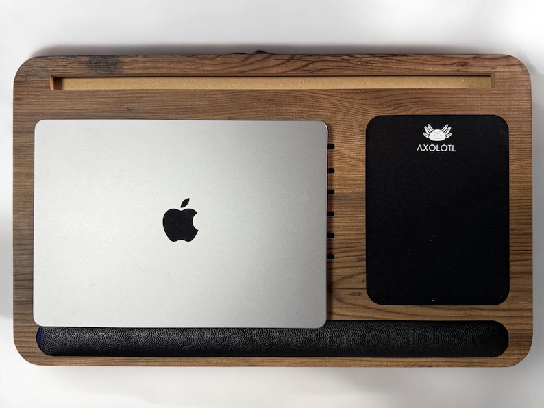 Może przedstawiać: Drewniany stojak na laptopa z czarnym sk&oacute;rzanym podparciem nadgarstka, srebrny laptop z logo Apple i czarny notes z napisem "AXOLOTL" nadrukowanym na biało.