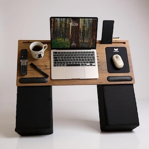 May include: A wooden laptop desk with a built-in mouse pad, wrist rest, and phone holder. The desk holds a laptop, mouse, and a smartphone. A coffee mug, remote control, and pen are also on the desk. The desk is supported by two black cushions.