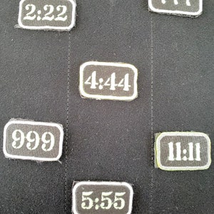 Op de afbeelding: Zes zwarte rechthoekige pleisters met witte cijfers op een zwarte achtergrond. De cijfers zijn 2:22, 4:44, 7:77, 9:99, 11:11 en 5:55.